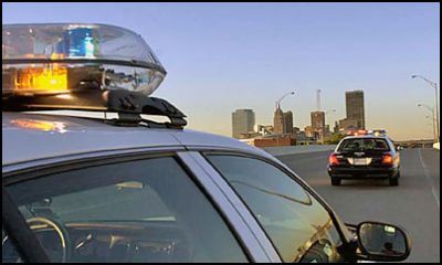 San Antonio police car with flashing lights on the highway, city skyline in background.