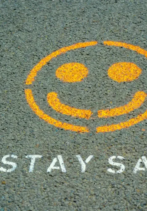 Pictured on asphalt is a smiley face in yellow chalk paint with the words "Stay Safe" in white chalk paint.